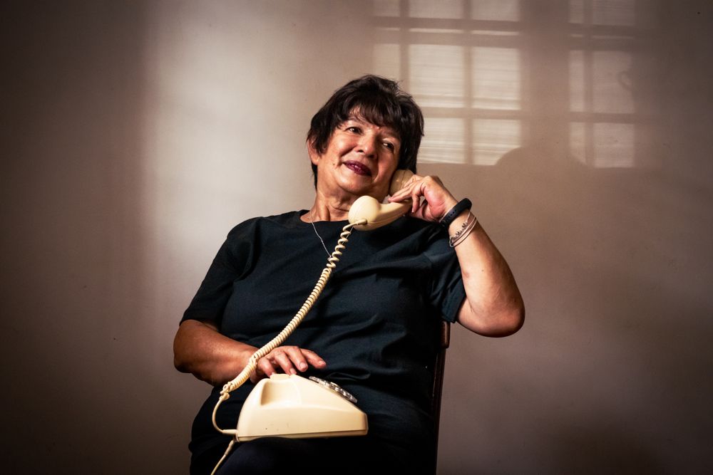 Une dame âgée souriante au téléphone fixe, assise dans une pièce lumineuse près d'une fenêtre
